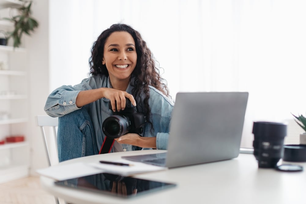 Happy photographer woman holding photo camera using laptop sitting